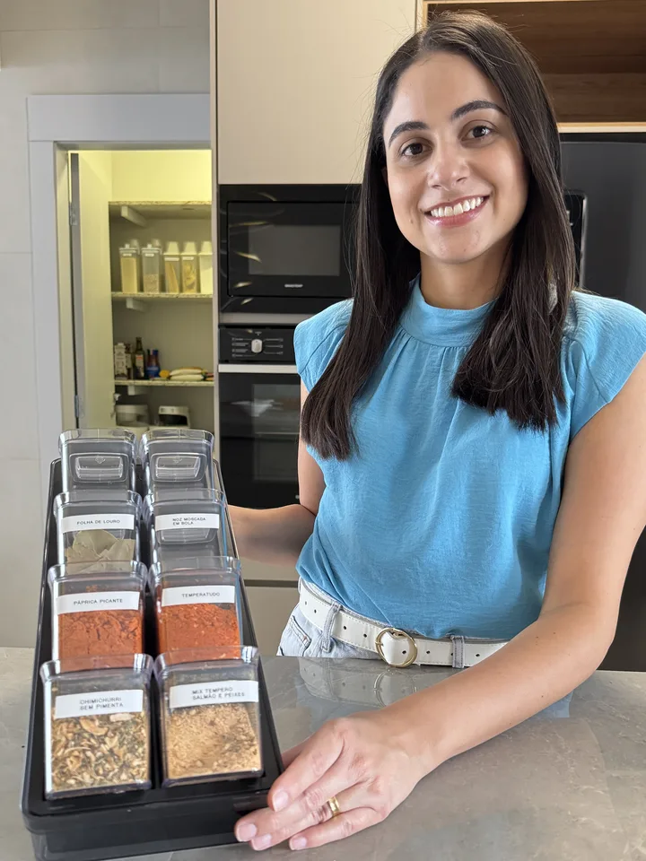 Mari ao lado da bancada da cozinha, com a bandeja de temperos em potes iguais e etiqueta em cada um